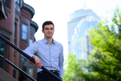 Vincent Rollet standing outside in Boston. An out-of-focus brownstone is visible to the left, with the hazy Prudential Center behind him.