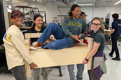 Four students carry a wooden bench they built while another student reclines atop it.