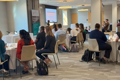More than 20 people sit at round tables in a conference room watching a presenter speak to slides projected on a screen.