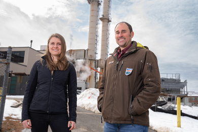 Jean Sack and Chris Lashway pose for a photo outside on a winter day, with a power plant in the background.