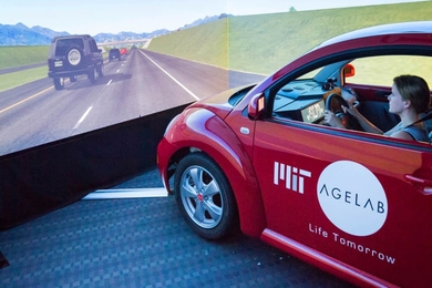 A woman steers a red coupe bearing the MIT AgeLab logo through a highway scene projected on three walls 