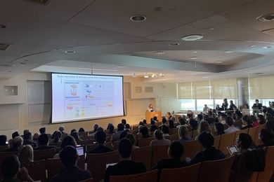 A wide shot of Singleton Auditorium in MIT's Building 46 shows an audience that fills the seats while a speaker at the front of the room presents an Alzheimer's research slide on a large screen