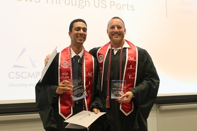 Yassine Lahlou-Kamal and Kevin Power in graduation regalia including "SCM" stoles, pose together holding clear glass awards