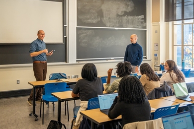 Robin Wolfe Scheffler and Mark Bathe stand in front of a classroom. Scheffler is speaking.