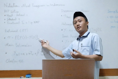 Munip Utama stands before a whiteboard, gesturing. A laptop sits open on the table in front of him.