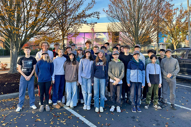 About 30 high school students and a teacher stand in front of a Boeing factory on a fall day
