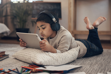 Young boy lying on the floor, reading a tablet while wearing headphones