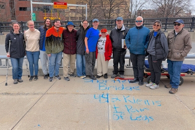 12 people stand together at the end of a bridge with the writing, “Shortfellow Bridge - 442 Kleins + pair of legs” in chalk on the sidewalk.