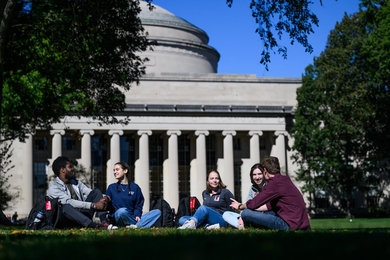 Four students sit on a grass courtyard on a sunny, early fall day. The MIT dome is in the background.