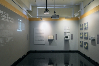 Wide view of a modest-sized room featuring wall-mounted prints, archival materials, digital displays, and geometric compositions