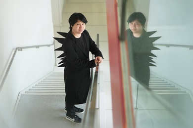 Pat Pataranutaporn stands on a staircase wearing a black coat with dramatic fabric spikes on his back, his reflection visible in a glass panel beside him.