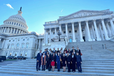 26 people in business attire pose standing on the steps of the U.S. Capitol building.