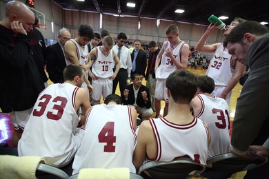 About nine members of MIT's men's basketball team huddle around with others during game