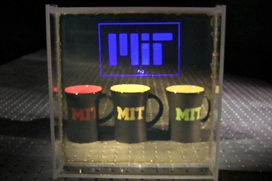A clear screen with "MIT" on it stands before three coffee mugs that also say "MIT" on them