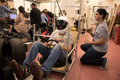 MIT senior Brian Wanek wears a racing helmet inside a prototype of what will be the driver’s seat in the 2017 MIT Formula SAE car as sophomore Wasay Anwer measures the frame.
