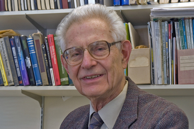 Headshot photo of John Dugundji sitting in an office in front of a bookshelf