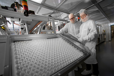 Photo of two technicians covered in white protective coveralls examining a tray of hundreds of little glass vials.