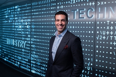 Portrait photo of Blade Kotelly standing before a background of computer code. He’s wearing a nice blazer and smiling.