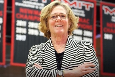 Photo of Julie Soriero standing with her arms folded, in front of championship banners hanging in an MIT gym