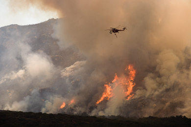 Photo of a helicopter flying over a wildfire on a hillside. You can see orange flames in the tree line and large clouds of white and grey smoke surrounding the area.