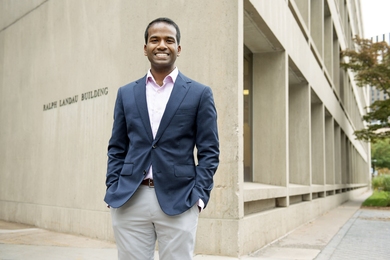 Karthish Manthiram profile photo. He is wearing a blazer and standing with his hands in his pockets in front of the Ralph Landau Building at MIT