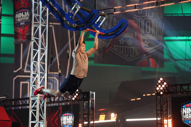 TV screen grab of a student on a green-lit obstacle course, swinging his legs backwards while holding onto a trapeze-style hanging bar in preparation for making a leap.