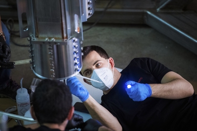 Photo of a young man wearing a white facemask and blue safety gloves, adjusting part of the experiment, holding a small flashlight while adjusting something on a metal cylinder, less than a foot wide and punctuated with dozens of small holes