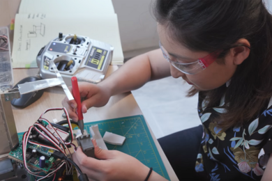 Photo of Julianna Rodriguez works on a robot on a kitchen table. She is holding an instrument that looks like a pen, manipulating a small piece of equipment with colorful wires popping out of it.