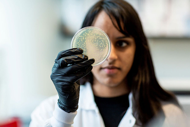 Closeup photo of a woman wearing a white lab coat and black safety gloves, holding a petri dish in front of her face