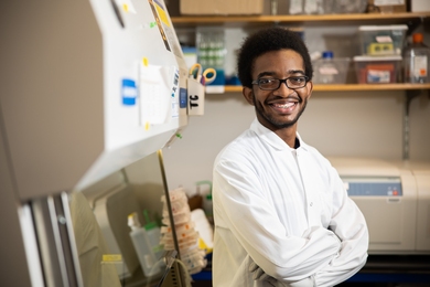 Photo of Desmond Edwards in a lab coat, standing with crossed arms in front of a hood in the lab.