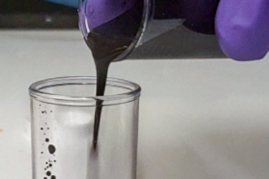 Gloved fingers pouring a black liquid from a test tube into a beaker