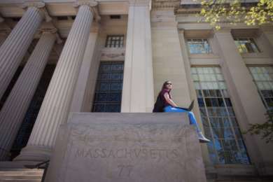 A student sits with a laptop open on a stone slab in front of two MIT buildings. The slab says "Massacvsetts 77"