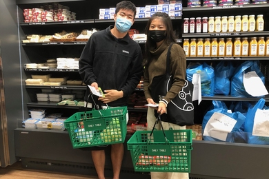 Photo of two students at a supermarket, posing with a basket of grocery items.
