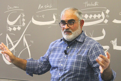 Hajj Wafaa standing in front of blackboard showing samples of Arabic calligraphy