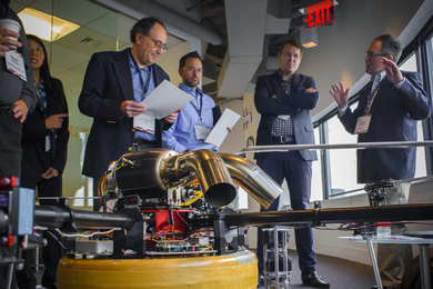 Six individuals stand around a small piece of equipment with metal tubes and wires.