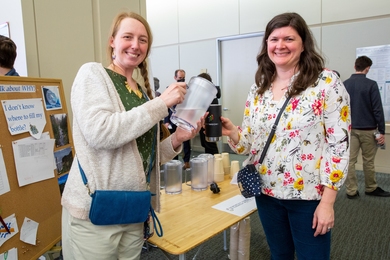Photo of Theresa Werth and Dorothy Hanna standing at a table with one pouring a pitcher of water into a mug. A bulletin board next to them has pictures of natural water scenes and the words "I don't know where to fill my bottle?"