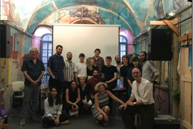 Photo of Mark Jarzombek posing with 16 students in a room with a colorfully painted arched ceiling. They are all posing in front of a pull-down media display screen.
