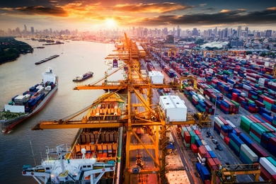 Photo of a busy port with ships being loaded and unloaded and thousands of containers waiting to be moved as the sun sets in the background.