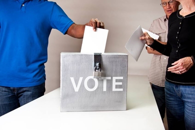 Photo of a box that says "vote," into which a person is placing a ballot, while two others wait their turn