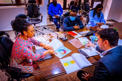 Photo of four people sitting around a table in a conference room, with two others seated behind them, looking on