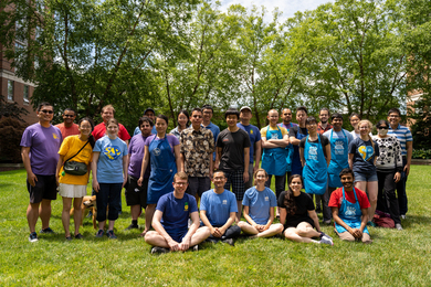 Several dozen people dressed in shorts and t-shirts pose for a group photo on a grassy area on a sunny day