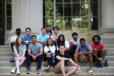 13 students pose for a group photo, sitting together on the steps of MIT Building 10, with columns and windows behind them.