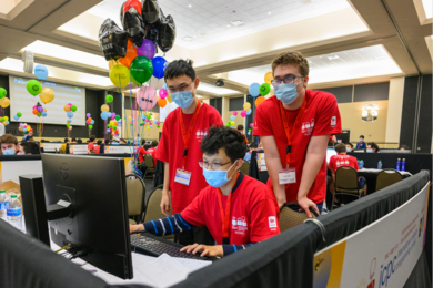Sharing a single computer in a room filled with similar teams of coders, three students in matching orange T-shirts and face masks work on their coding solutions.