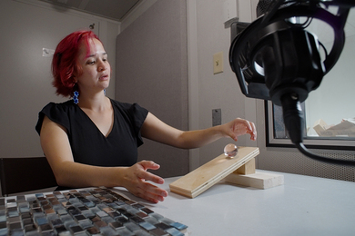 Fernanda De La Torre rolls a ball down a small wooden ramp on a table. A microphone hangs in the foreground, while a square foot sample of small glass tiles is at left.