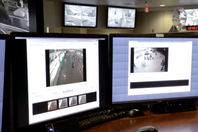 In the foreground, two desktop monitors display surveillance videos of a subway and a street scene. Two more monitors are mounted near the ceiling in the background