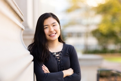 Sherry Nyeo leans against the outside of a stone building, with green trees in the background
