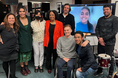 Eight smiling people in group next to one person joining virtually via a monitor