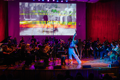 A dancer reaches their arm up in an on-stage performance in front of an orchestra and a large screen, which shows an abstract collage of people and colorful shapes