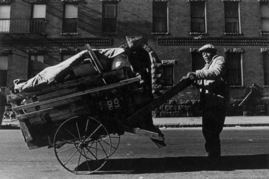 1960s black-and-white photo of a man pushing a two-wheeled cart on the street in front of a brick building