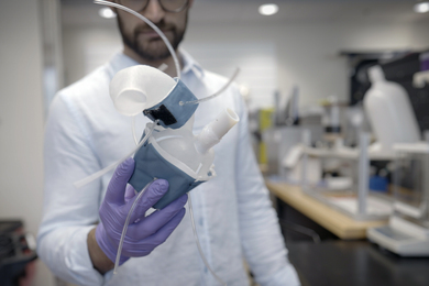 A person in a lab coat holds a blue and white 3d-printed heart replica, a soft and flexible shell with fabricated sleeves, in their gloved hand.